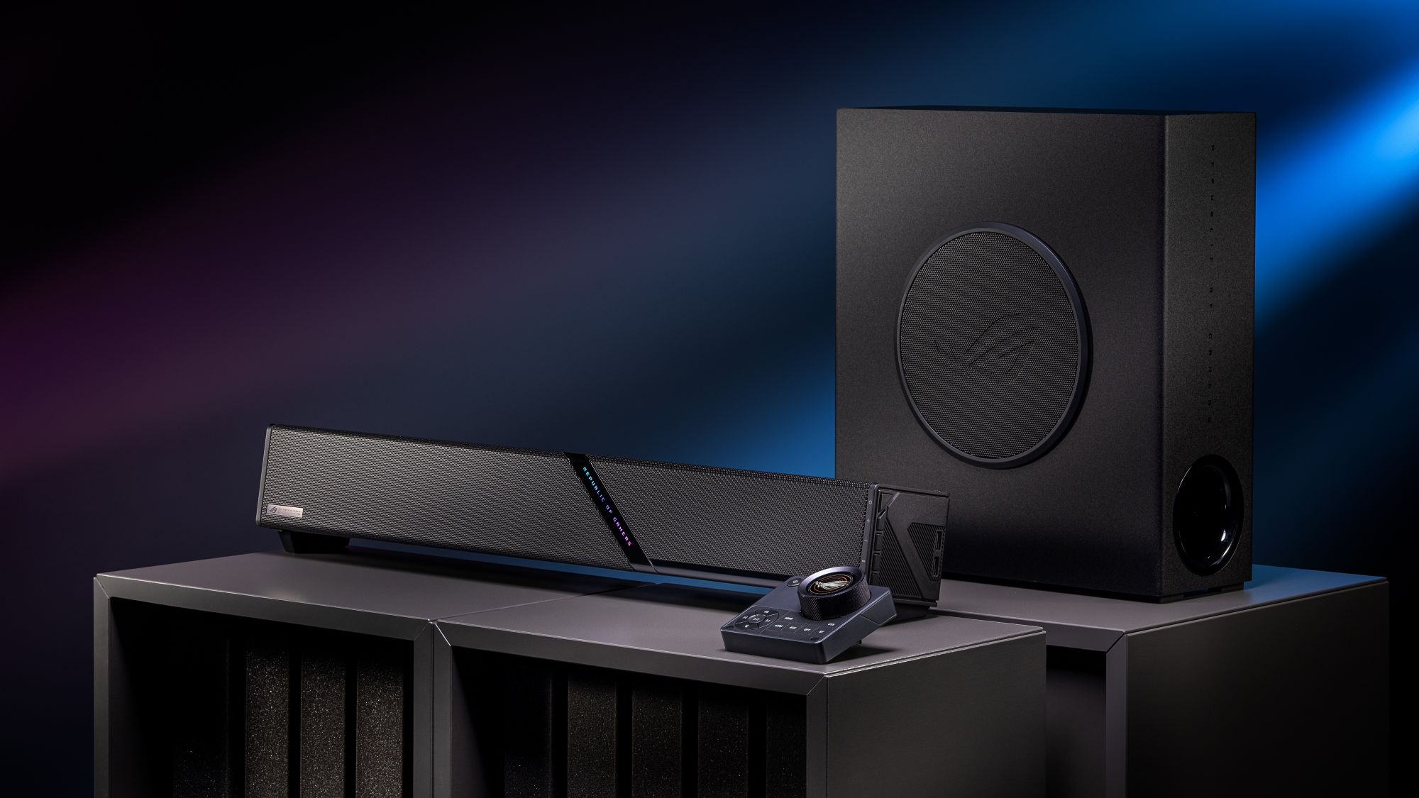 The ROG Gjallar gaming soundbar, control hub and wireless subwoofer sitting on black stands against a blue background.
