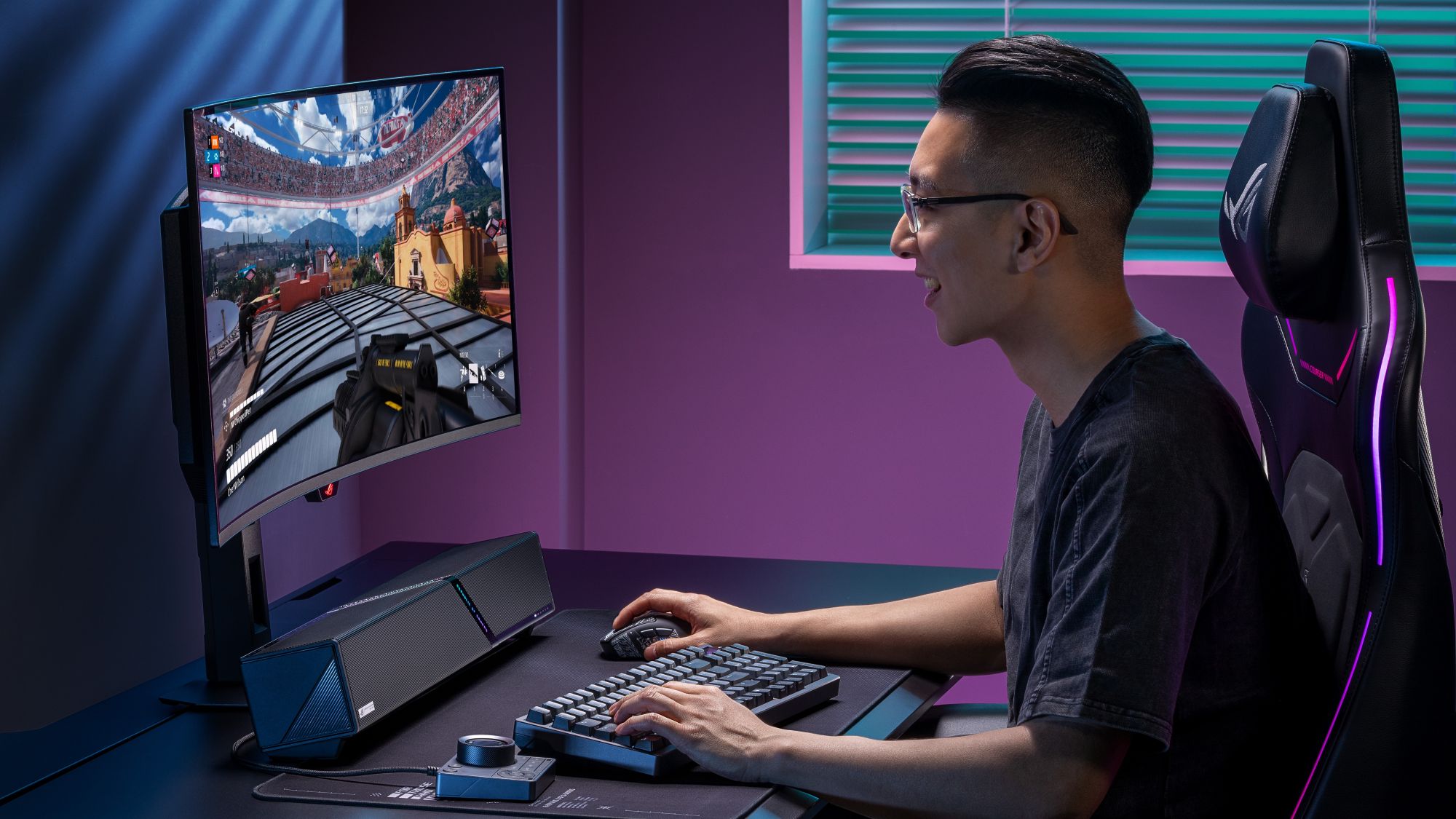 A man plays a game on his PC while the ROG Gjallar gaming soundbar sits on his black desk surrounded by ROG gear.